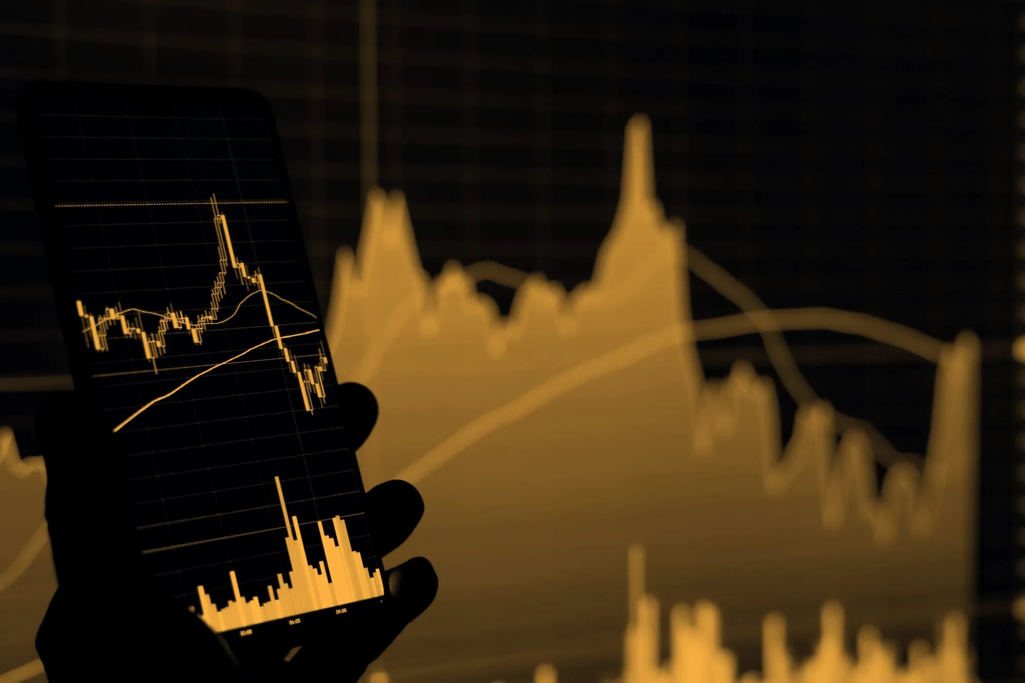 A phone held up to a wall of candlestick charts, the phone's own chart lit against the dark.